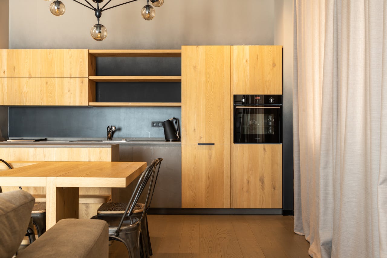 Wooden table with chairs placed in stylish kitchen with cupboards and modern appliances in apartment with curtains on window and couch